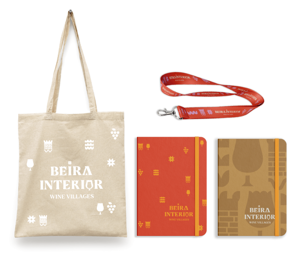 Conjunto de artigos de marketing de Vinhos da Beira Interior: tote bag branca, lanyard vermelho e dois cadernos de anotações com o logótipo 'BEIRA INTERIOR WINE VILLAGES'