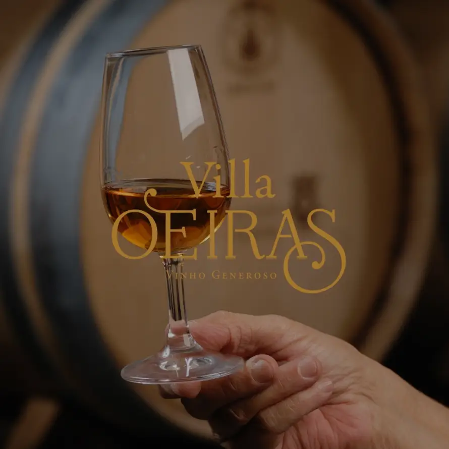 Mão a segurar um copo de vinho generoso Villa Oeiras com um barril de madeira ao fundo.