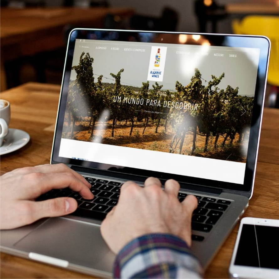 Pessoa a consultar o website oficial Algarve Wines "Um Mundo para Descobrir" num MacBook, com um vinha algarvia no ecrã